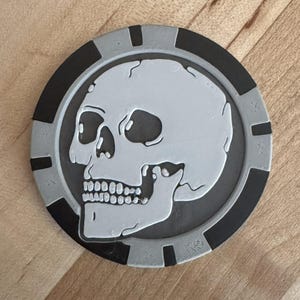 May include: A round, gray and black skull-themed coaster. The skull is white with black eye sockets and a detailed jawline. The coaster has a gray border with black rectangular accents.