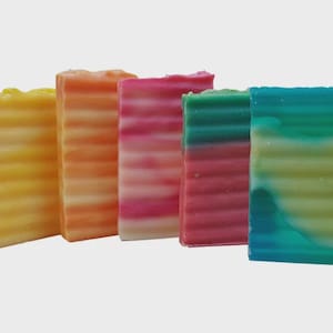 Könnte beinhalten: Fünf bunte handgemachte Seifenstücke. Die Seifen sind nebeneinander angeordnet, jede mit einem einzigartigen Farbverlauf. Zu den Farben gehören Gelb, Orange, Rosa, Grün und Blau. Die Seifen haben eine strukturierte, geriffelte Oberfläche.