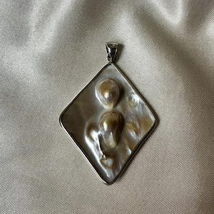 May include: A silver pendant with a diamond-shaped frame. The pendant features two irregularly shaped, golden-brown pearls set against a pearlescent background. The pendant has a small loop at the top for a chain.