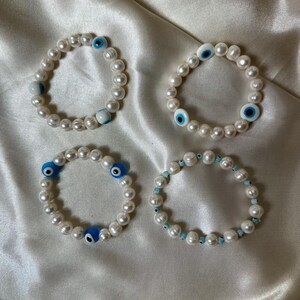 May include: Four pearl bracelets with blue evil eye and light blue bead accents. The bracelets are made of white pearls and are arranged on a white satin surface. The evil eye beads are blue and white.