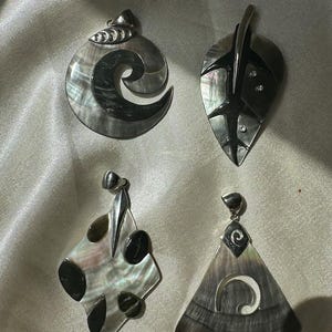 Könnte beinhalten: Vier schillernde Muschel-Anhänger in verschiedenen Formen: eine Spirale, ein Blatt, eine Raute und ein Dreieck. Jeder Anhänger hat eine silberne Öse zum Aufhängen. Die Muschelfarben reichen von Grau bis Schwarz mit schillernden Akzenten.