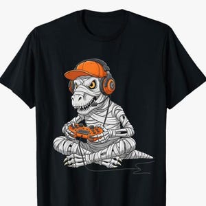 Puede incluir: Camiseta negra con un diseño de dinosaurio momia de dibujos animados. El dinosaurio lleva una gorra y auriculares naranjas y sostiene un mando de juego naranja. El dinosaurio está envuelto en vendas blancas y tiene ojos amarillos.