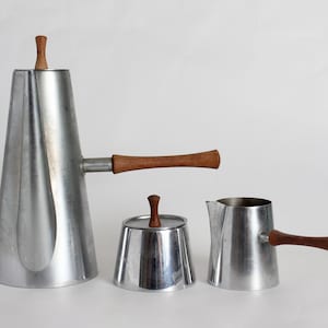 Könnte beinhalten: Ein silbernes Kaffeekännchen, Milchkännchen und Zuckerdose mit Holzgriffen. Die Kaffeekanne hat eine hohe, konische Form mit einem Ausguss und einem Deckel. Das Milchkännchen und die Zuckerdose sind beide rund mit Deckeln.
