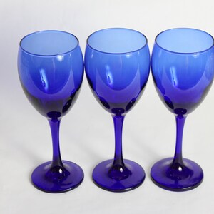 Peut inclure: Trois verres &agrave; vin bleu cobalt avec une base claire l&eacute;g&egrave;rement teint&eacute;e. Les verres sont dispos&eacute;s en rang&eacute;e sur une surface blanche.