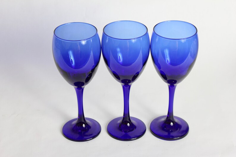 Peut inclure: Trois verres &agrave; vin bleu cobalt avec une base claire l&eacute;g&egrave;rement teint&eacute;e. Les verres sont dispos&eacute;s en rang&eacute;e sur une surface blanche.