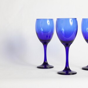 Peut inclure: Trois verres &agrave; vin bleu cobalt avec un pied transparent sur une surface blanche.
