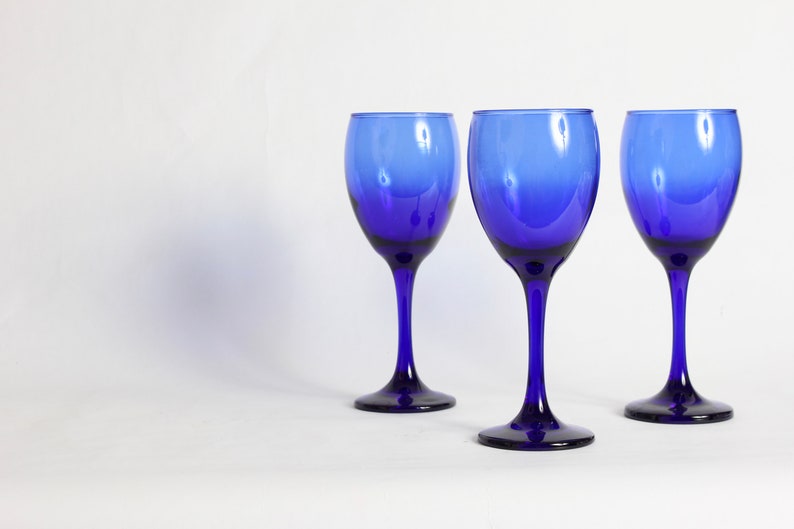 Peut inclure: Trois verres &agrave; vin bleu cobalt avec un pied transparent sur une surface blanche.