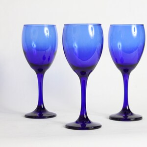 Peut inclure: Trois verres &agrave; vin bleu cobalt avec une base claire l&eacute;g&egrave;rement teint&eacute;e. Les verres sont dispos&eacute;s en rang&eacute;e sur une surface blanche.