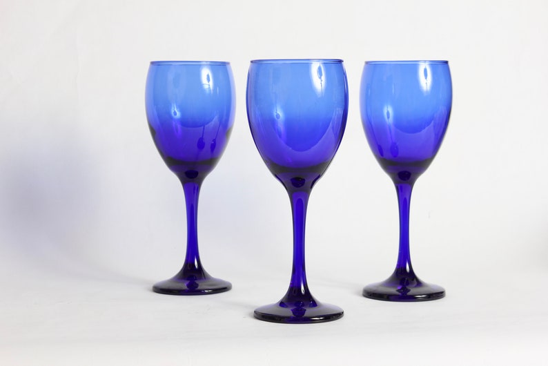 Peut inclure: Trois verres &agrave; vin bleu cobalt avec une base claire l&eacute;g&egrave;rement teint&eacute;e. Les verres sont dispos&eacute;s en rang&eacute;e sur une surface blanche.