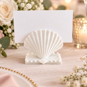 May include: A white seashell-shaped place card holder featuring a rectangular base adorned with small white pearls. The holder supports a blank rectangular white card, providing a clean and structured display for table settings or event signage.