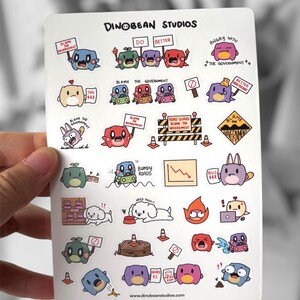 May include: A sticker sheet featuring various cartoon characters with expressions and signs. The characters are in different colors, some holding signs that say "Blame the Government" and "Do Better". The sheet includes the text "DinoBean Studios".