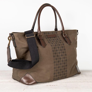 Puede incluir: Bolso tote verde oliva con detalles en cuero marrón. Cuenta con una correa de hombro negra y asas de cuero marrón. El bolso tiene un patrón decorativo en el centro y esquinas de cuero marrón.