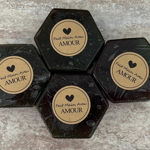 May include: Four dark, hexagonal soaps are arranged in a group. Each soap has a round, tan label with a black heart and the words "Fait Main Avec AMOUR" in a circular arrangement. The soaps appear to be wrapped.