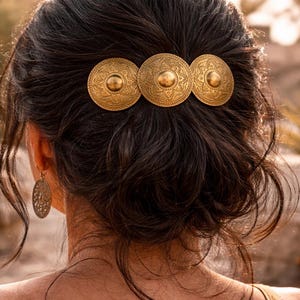 May include: A woman's dark hair is styled with a gold hair clip. The clip features three circular, ornate medallions with embossed patterns and a central raised detail. The hair accessory adds a touch of elegance.