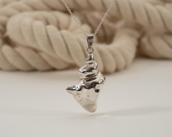 May include: A silver-colored pendant necklace featuring a seashell design. The pendant is suspended from a delicate chain. The background includes a thick, off-white rope and a white surface.