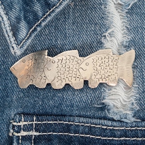 May include: A silver-toned brooch featuring three stylized fish, each with detailed scale patterns and eye markings. The brooch is pinned to a denim jacket, showcasing its design and texture. The fish are connected, creating a unique accessory.
