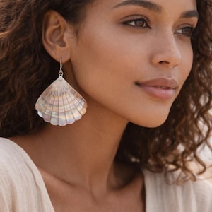 May include: A woman wearing a large, iridescent seashell earring. The earring is a fan shape with a silver hook. The woman has brown curly hair and is wearing a cream-colored top. The background is blurred, suggesting a beach setting.