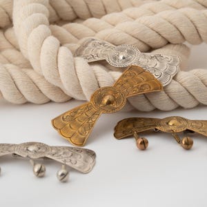 May include: Four decorative metal hair barrettes in silver and gold tones. The barrettes have intricate engraved designs and some feature small bell-shaped accents. A thick, off-white rope is in the background.