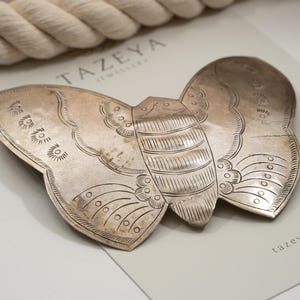 May include: A silver-toned butterfly-shaped brooch with intricate etched details. The brooch features a detailed body and wings, resting on a light gray surface with the brand name "TAZEYA" and website address.