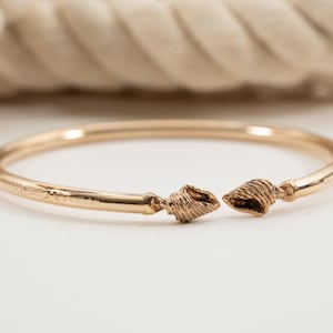 May include: A gold-colored bangle bracelet with seashell accents. The bracelet is open at the front, with a seashell design on each end. The bracelet is made of a shiny, metallic material.