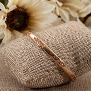 May include: A gold-colored bangle bracelet with an intricate floral design. The bracelet is resting on a small, square, tan pillow. The background features cream-colored flowers.