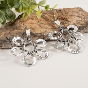 May include: A pair of silver-toned metal earrings. Each earring features a looped, abstract floral design. The metal has a hammered texture, and the earrings hang from simple ear wires. The earrings are displayed on a natural stone surface.