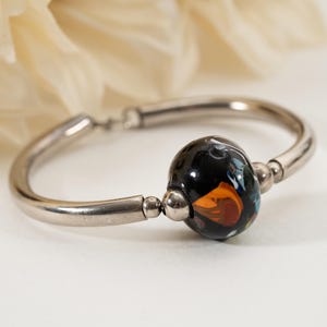 May include: A silver-toned bangle bracelet with a central, large, round bead. The bead is primarily black with swirls of orange, white, and blue. The bracelet is on a white surface with a blurred, off-white background.