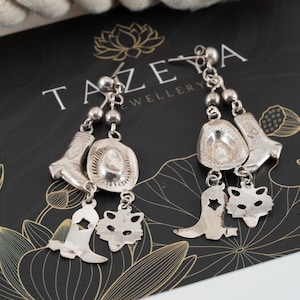 May include: A pair of silver-toned dangle earrings featuring charms shaped like cowboy boots, cowboy hats, and fox heads. Each earring is suspended from a small silver ball stud and includes multiple dangling elements attached by small metal rings.