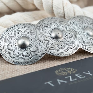 May include: Three silver-colored, circular decorative elements with embossed floral patterns and a central raised stud. The elements are arranged in a row, with a textured rope in the background. The brand name "TAZEYA JEWELLERY" is visible.
