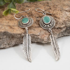 May include: A pair of silver earrings with a malachite stone center and a feather pendant. The earrings have a detailed, ornate design around the stone and on the feather. The malachite stone is a vibrant green color.