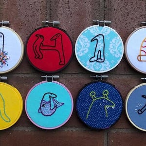 Peut inclure: Huit cerceaux brodés avec des motifs de créatures fantaisistes. Chaque cerceau présente un fond de couleur différente, notamment blanc, rouge, bleu et jaune. Les créatures sont soulignées en noir et cousues avec diverses couleurs, créant un affichage ludique et artistique.