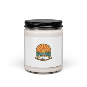 Puede incluir: Un tarro de vela blanco con tapa negra. La etiqueta presenta un diseño retro con un sol, palmeras y una furgoneta en una carretera, con el texto "ENDLESS SUMMER ROAD TRIP".
