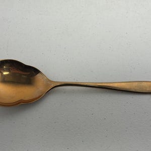 May include: A gold-colored spoon with a scalloped bowl and a slender handle. The spoon is made of metal and has a polished finish, reflecting light. The spoon is designed for serving.