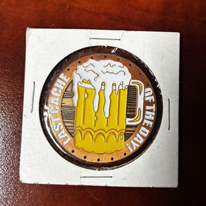 以下が含まれることがあります： 黄色いビールジョッキとあふれる白い泡が特徴の、ビールをテーマにした丸いピン。 ピンは茶色の背景に「LAST CACHE OF THE DAY！」の文字が円形に配置されています。 白い厚紙ホルダーに入っています。