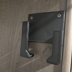 May include: A black, 3D-printed wall mount with two angled arms and a rectangular backplate. The mount is attached to a wooden surface with two screws. The design appears to be for holding an item.