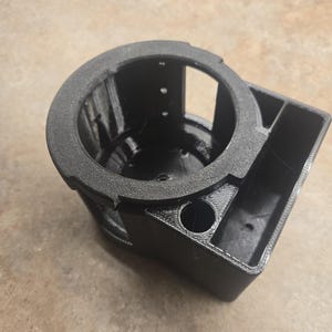 May include: A black, 3D-printed car cup holder with a textured rim. The holder features a circular cup space and a rectangular compartment. The design includes several small holes and a rounded corner.