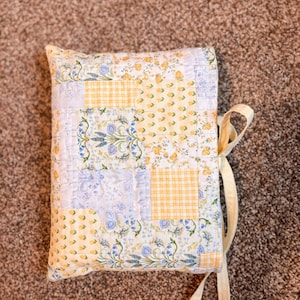 Puede incluir: Una funda de libro acolchada con diseño de patchwork. Presenta cuadrados de tela floral amarilla, floral azul y cuadros amarillos. Se adjunta una cinta color crema para el cierre. La estética general es vintage y campestre.