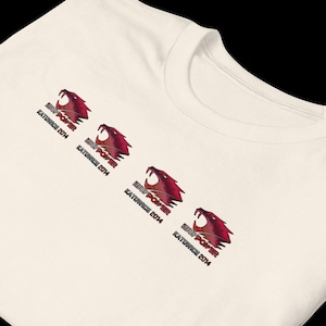 May include: Cream-colored t-shirt featuring four red and black snarling cougar head graphics. Each graphic is accompanied by the text "IBUYPOWER KATOWICE 2014" in black. The shirt is made of a soft, comfortable material.
