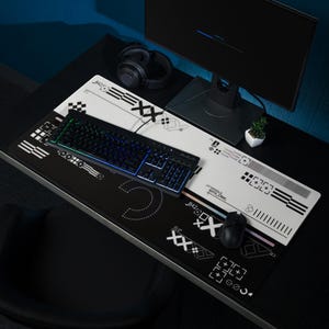 May include: A black and white gaming mouse pad with a keyboard and mouse. The pad has a geometric design with the text "HANDLE WITH CARE". A black keyboard with illuminated keys sits on the pad. A computer monitor and headphones are also visible.