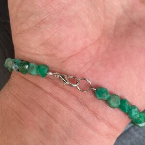 May include: A bracelet composed of small, faceted, vibrant green gemstone beads. The piece features a silver-toned metal lobster claw clasp and a circular jump ring connector, securing the strand of irregularly shaped, polished green stones.