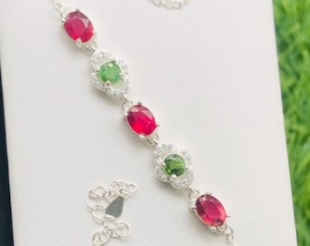 May include: A silver bracelet featuring oval-cut red and green gemstones. The bracelet has a delicate chain design with a lobster clasp closure. The gemstones are set in a floral design with small, clear accent stones.