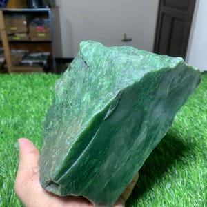 Puede incluir: Un trozo grande y bruto de jade nefrita de color verde intenso con una textura superficial natural e irregular. La piedra muestra tonos variados de verde bosque y esmeralda con estructuras cristalinas visibles y facetas lisas.