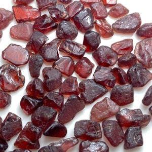 Puede incluir: Una colección de piedras preciosas de granate en bruto, sueltas, con formas angulares irregulares y tonos variados de rojo intenso y burdeos. Cada piedra muestra una calidad de semitranslúcida a opaca con una textura natural y bordes facetados.
