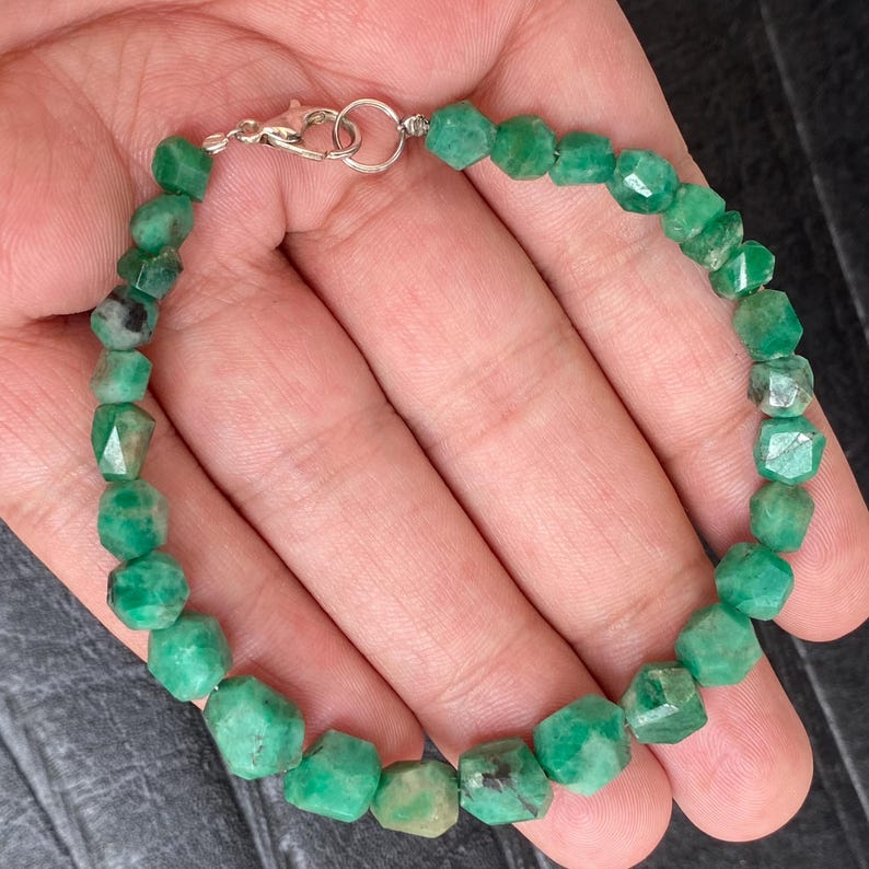 May include: A beaded bracelet featuring faceted, semi-translucent green emerald stones in varying shades of forest and mint green. The bracelet is secured with a silver-toned metal lobster claw clasp and a circular jump ring connector.
