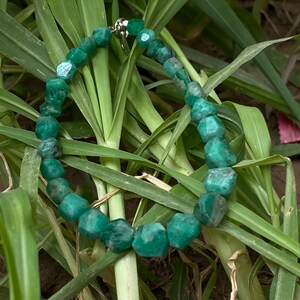 May include: A necklace composed of faceted, deep green gemstone beads strung together in a circular arrangement. The beads exhibit natural variations in color and texture, and the strand is secured with a small, metallic silver-toned clasp.