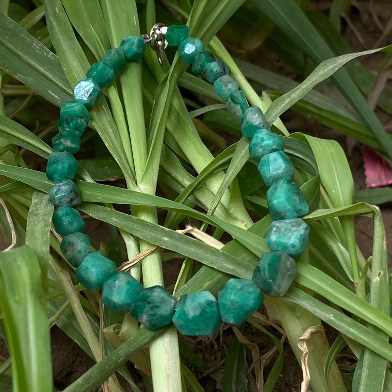 May include: A necklace composed of faceted, deep green gemstone beads strung together in a circular arrangement. The beads exhibit natural variations in color and texture, and the strand is secured with a small, metallic silver-toned clasp.