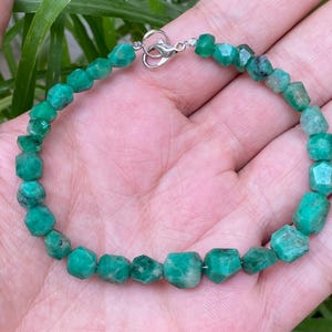 May include: A bracelet composed of small, faceted, vibrant green emerald beads strung together. The piece features a polished silver-toned metal lobster claw clasp that connects the two ends of the beaded strand securely.