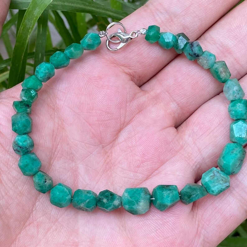May include: A bracelet composed of small, faceted, vibrant green emerald beads strung together. The piece features a polished silver-toned metal lobster claw clasp that connects the two ends of the beaded strand securely.