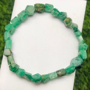 May include: A green emerald bracelet. The bracelet is made of faceted, irregular emerald beads. The beads are a vibrant green color with some brown inclusions. The bracelet is displayed on a white surface with a green background.