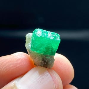 May include: A raw, vibrant green emerald crystal with a distinct cubic shape, attached to a rough, light-colored mineral matrix. The gemstone displays a translucent quality and a bright, saturated emerald green hue, showcasing natural crystalline facets.